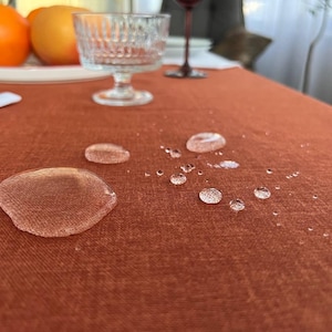 May include: Close-up of a rust-coloured tablecloth with multiple water droplets on the surface. The droplets are round and clear, indicating that the tablecloth is water-resistant.