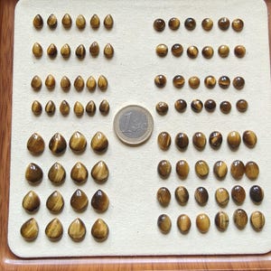 May include: A collection of polished tiger's eye gemstones in various shapes and sizes, arranged on a light-colored surface. The gemstones display a range of brown and gold hues, with a coin for size comparison. The gemstones are teardrop, oval, and round shaped.