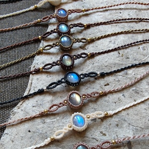 Può includere: Una collezione di braccialetti in macramè con pietre di labradorite ovali dai riflessi blu. Ogni braccialetto è realizzato con un cordino sottile in varie tonalità della terra, tra cui marrone, nero, beige e crema, con piccole perline metalliche.