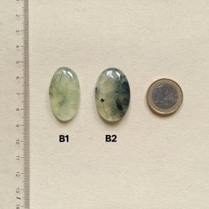 May include: Two oval-shaped prehnite gemstones, labeled B1 and B2, next to a Euro coin for size comparison. The gemstones are pale green with darker inclusions. A ruler is on the left side of the image.