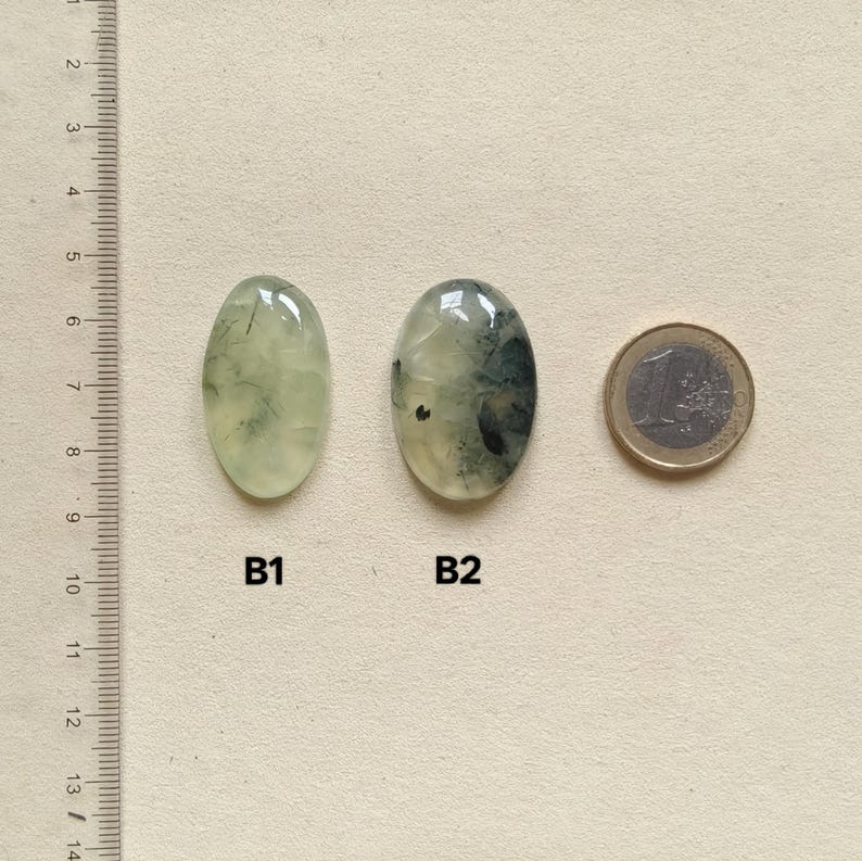 May include: Two oval-shaped prehnite gemstones, labeled B1 and B2, next to a Euro coin for size comparison. The gemstones are pale green with darker inclusions. A ruler is on the left side of the image.