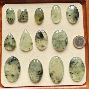 May include: A collection of polished prehnite gemstones in various shapes, including oval, teardrop, and rectangular. The translucent stones have a pale green hue with dark inclusions, displayed in a wooden tray. A Euro coin is included for size reference.