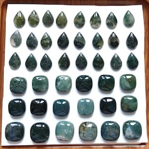 Puede incluir: Una variedad de piedras preciosas de ágata musgosa en varias formas, incluyendo lágrimas y cuadrados, exhibidas sobre una superficie blanca. Las piedras varían en color desde verde claro hasta verde bosque oscuro, con patrones únicos. Las piedras están dispuestas ordenadamente.