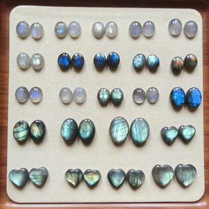 May include: A selection of polished labradorite gemstones in various shapes, including oval and heart-shaped stones. The stones display iridescent blue, green, and grey hues, arranged on a beige surface.