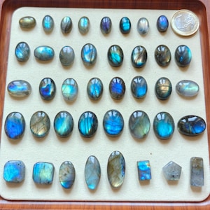 Pode incluir: Uma coleção de pedras preciosas de labradorite polidas em várias formas e tamanhos, incluindo ovais e retangulares. As pedras exibem flashes iridescentes de azul, verde e dourado. Uma moeda de euro está incluída para comparação de tamanho.