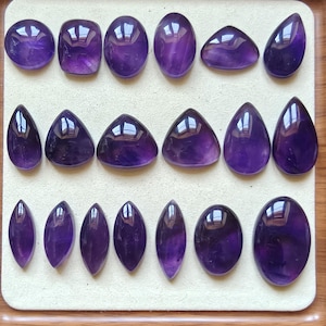 Könnte beinhalten: Eine Sammlung von achtzehn polierten, tiefvioletten Amethyst-Cabochons, angeordnet in drei Reihen. Die Edelsteine haben verschiedene Formen, darunter runde, ovale, tropfenförmige, marquise- und dreieckige Schliffe mit glatter Oberfläche.
