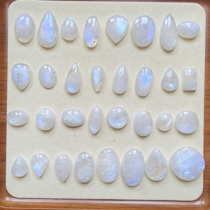 Peut inclure: Une collection de trente et un cabochons de pierre de lune polis disposés en quatre rangées sur un présentoir beige clair. Les pierres précieuses présentent diverses formes, notamment ovale, poire, marquise et rectangulaire, toutes blanches.