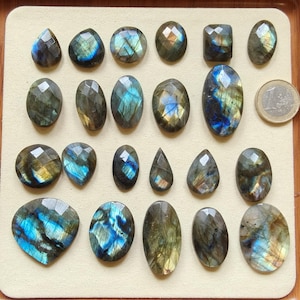Könnte beinhalten: Eine Sammlung facettierter Labradorit-Edelsteine in verschiedenen Formen, darunter oval, tropfenförmig und rund. Die Steine zeigen irisierende blaue, grüne und goldene Blitze. Eine Euro-Münze ist zum Größenvergleich enthalten.