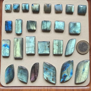 Può includere: Una collezione di pietre preziose di labradorite lucidata in varie forme, tra cui quadrati, rettangoli e design a forma libera. Le pietre mostrano lampi iridescenti blu, verdi e viola. Una moneta da euro è inclusa come riferimento delle dimensioni. Le pietre sono disposte su una superficie beige.