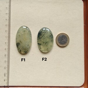 May include: Two oval-shaped prehnite gemstones with inclusions, labeled F1 and F2, are displayed next to a Euro coin for size comparison. The gemstones are light green with dark green and black inclusions. A ruler is in the background.