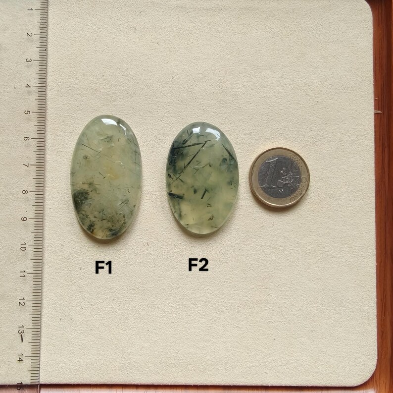 May include: Two oval-shaped prehnite gemstones with inclusions, labeled F1 and F2, are displayed next to a Euro coin for size comparison. The gemstones are light green with dark green and black inclusions. A ruler is in the background.