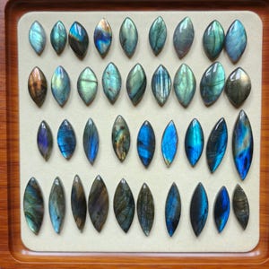 May include: A display of oval labradorite gemstones, exhibiting a spectrum of blues, greens, and golds. The stones are presented on a cream-coloured surface within a wooden tray, highlighting their iridescent nature.