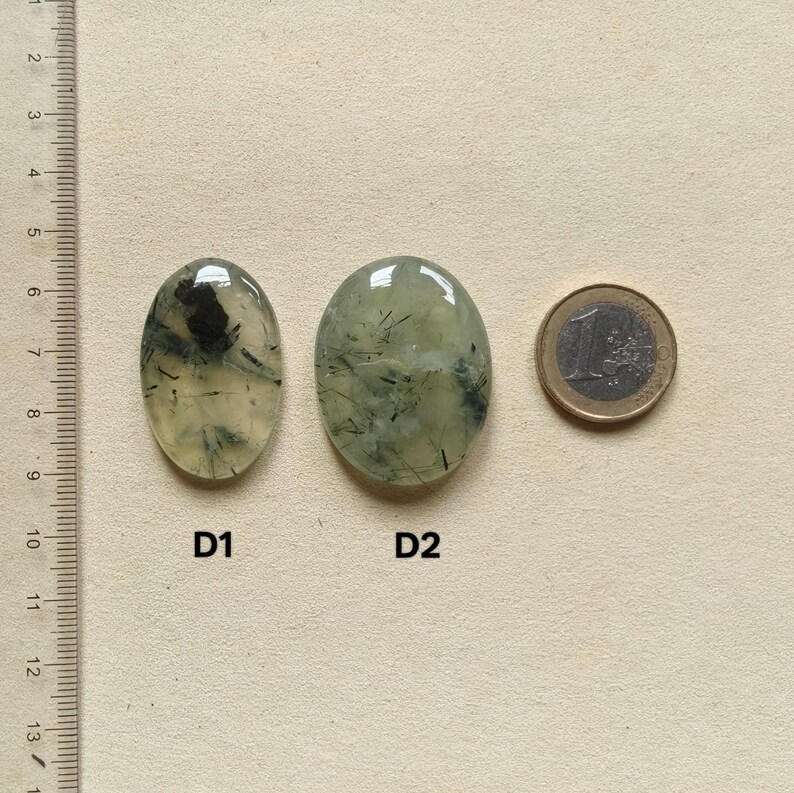 May include: Two oval-shaped prehnite gemstones with inclusions, labeled D1 and D2, next to a Euro coin for size comparison. The gemstones are translucent with green and black patterns. A ruler is on the left side of the image.