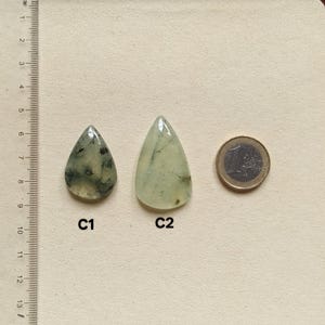 May include: Two teardrop-shaped prehnite gemstones, one with dark inclusions, the other with light inclusions, next to a Euro coin. The gemstones are light green and translucent. A ruler with centimeter markings is in the background.