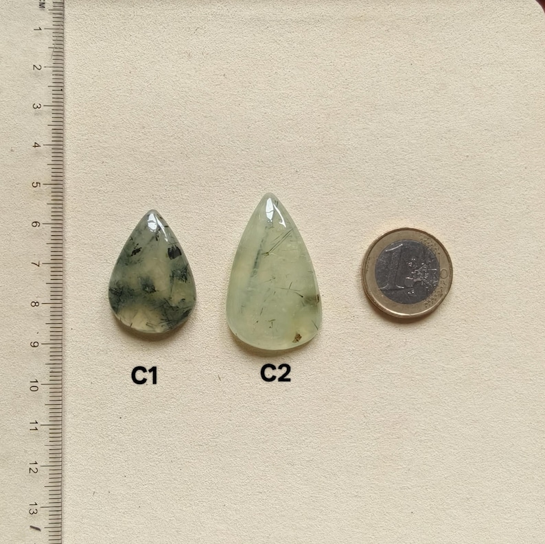 May include: Two teardrop-shaped prehnite gemstones, one with dark inclusions, the other with light inclusions, next to a Euro coin. The gemstones are light green and translucent. A ruler with centimeter markings is in the background.