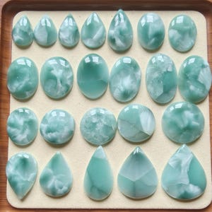 May include: A collection of twenty-two smooth, polished cabochons in varying shades of seafoam green and white. The stones are arranged in four rows, featuring a mix of teardrop and oval shapes with marbled, cloud-like patterns throughout.