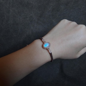 Könnte beinhalten: Handgefertigtes Makramee-Armband mit einem leuchtenden, ovalen Mondstein. Das Armband hat eine braune Kordel mit beigen Akzenten und kleinen goldfarbenen Perlen. Der Mondstein ist etwa 2 cm lang.