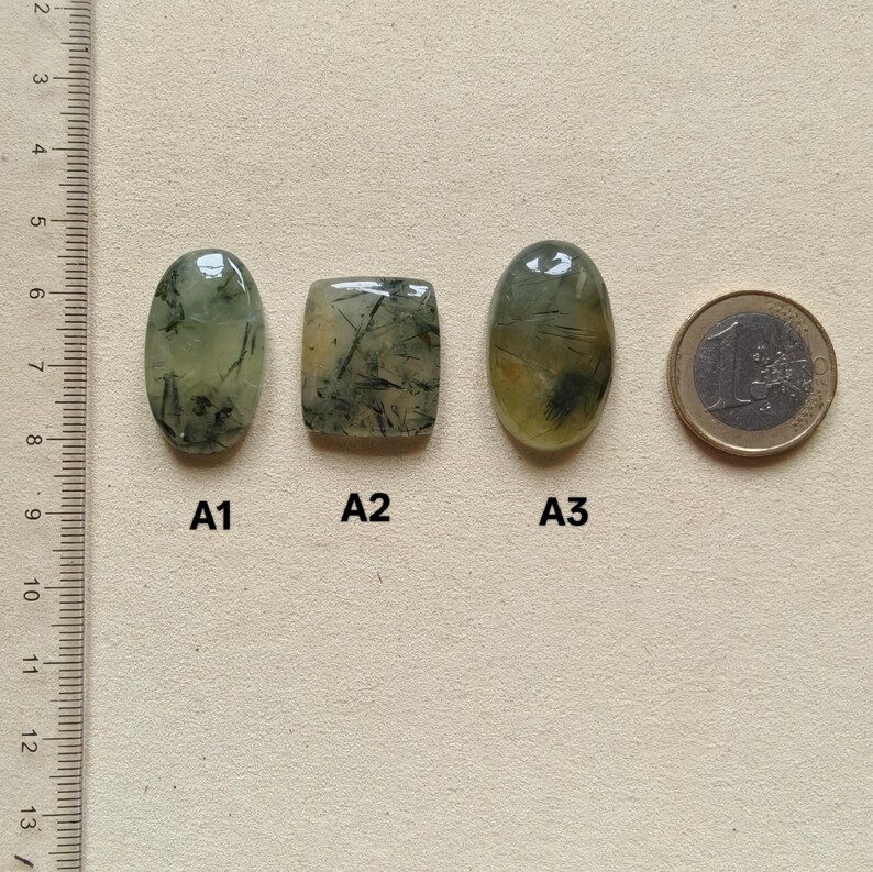 May include: Three polished gemstones with inclusions, labeled A1, A2, and A3, next to a Euro coin. The gemstones are oval and rectangular, with a translucent green hue and dark, needle-like inclusions. A ruler is in the background.