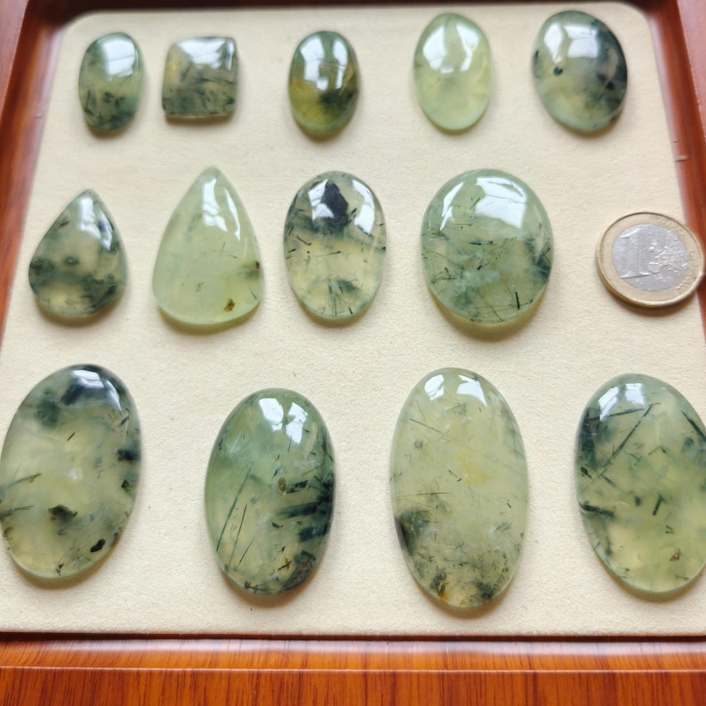 May include: A collection of polished prehnite gemstones in various shapes, including oval, teardrop, and square. The translucent stones display a pale green hue with dark inclusions, arranged in a wooden tray. A coin is included for size reference.