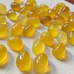 Può includere: Una collezione di pietre preziose lisce a forma ovale in varie tonalità di giallo e ambra. Le pietre lucide hanno una superficie lucida e riflettente e sono sparse su uno sfondo neutro. Le pietre preziose sembrano naturali, forse cabochon lucidi.