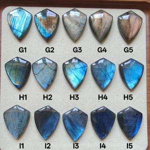 May include: A collection of fifteen shield-shaped labradorite pendants arranged in three rows of five, labelled G1 through G5, H1 through H5, and I1 through I5. Each polished gemstone displays unique iridescent blue, grey, and brown tones.
