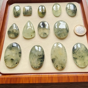 May include: A collection of polished prehnite gemstones in various shapes, including oval, teardrop, and rectangular. The translucent stones have a pale green hue with dark inclusions, displayed on a beige surface within a wooden tray. A Euro coin is also present.