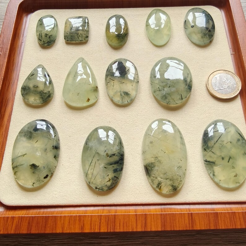 May include: A collection of polished prehnite gemstones in various shapes, including oval, teardrop, and rectangular. The translucent stones have a pale green hue with dark inclusions, displayed on a beige surface within a wooden tray. A Euro coin is also present.