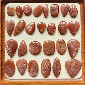 Pode incluir: Uma coleção de pedras preciosas de pedra do sol polidas em várias formas, incluindo ovais, em forma de gota e retangulares. As pedras têm um tom quente, avermelhado-alaranjado com inclusões brilhantes, exibidas sobre uma superfície bege numa bandeja de madeira.