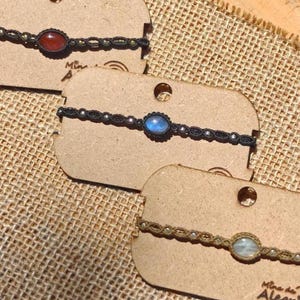 Può includere: Primo piano di tre braccialetti in macramè con pietre preziose ovali rosse, blu e bianche. I braccialetti sono esposti su piccole etichette di cartone marrone chiaro su uno sfondo beige strutturato. Ogni braccialetto ha una gemma unica.