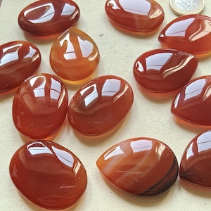 Può includere: Una collezione di pietre preziose di corniola lucidata in varie forme, tra cui ovali e a goccia. Le pietre mostrano sfumature di arancione e marrone rossiccio con una finitura lucida. Una moneta è visibile sullo sfondo.