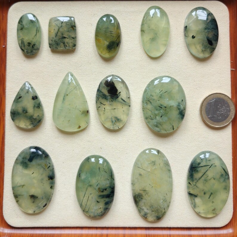 May include: A collection of polished prehnite gemstones in various shapes, including oval, teardrop, and rectangular. The translucent stones display a pale green hue with dark inclusions, arranged on a cream-colored surface. A Euro coin is included for size reference.