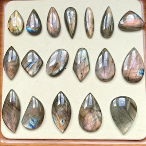 Könnte beinhalten: Eine Sammlung polierter Labradorit-Edelsteine in verschiedenen Formen, darunter Tropfen, Oval und Diamant. Die Steine zeigen schillernde Blitze in Blau, Gold und Lila innerhalb einer grauen und braunen Matrix. Die Edelsteine sind auf einer cremefarbenen Oberfläche angeordnet.
