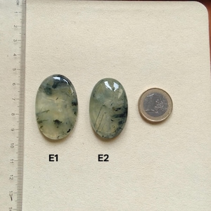 May include: Two oval-shaped, polished gemstones with green and black inclusions are displayed next to a Euro coin. The gemstones, labeled E1 and E2, have a translucent appearance. A ruler in centimeters is visible on the left side of the image.