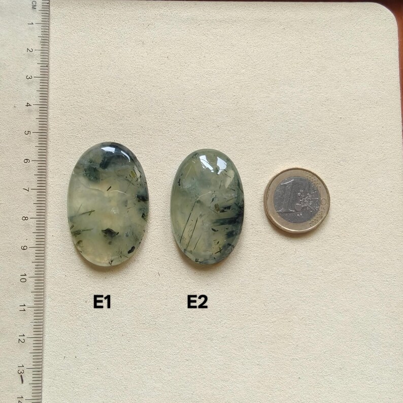 May include: Two oval-shaped, polished gemstones with green and black inclusions are displayed next to a Euro coin. The gemstones, labeled E1 and E2, have a translucent appearance. A ruler in centimeters is visible on the left side of the image.