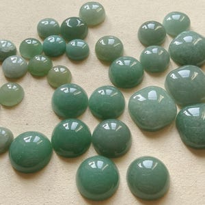 Könnte beinhalten: Eine Sammlung polierter grüner Edelsteine in verschiedenen Formen und Größen. Die Steine reichen von runden Cabochons bis hin zu unregelmäßigen, getrommelten Formen, alle mit einer glänzenden, reflektierenden Oberfläche. Die Farbe variiert von hellen bis zu tiefen Grüntönen.