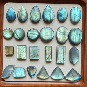 May include: A selection of polished labradorite gemstones in diverse shapes, including teardrops, ovals, circles, rectangles, and triangles. The stones exhibit iridescent blue, green, and gold colours, arranged on a beige surface within a wooden tray.