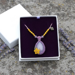 Puede incluir: Un collar de macramé hecho a mano con un colgante en forma de lágrima en una caja de regalo blanca. El colgante es lavanda con un acento floral amarillo. El collar tiene cordones amarillos y rosas con cuentas transparentes. Ramas de lavanda a los lados.