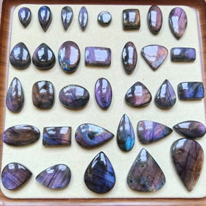 Può includere: Una collezione di pietre preziose in labradorite lucida di varie forme, tra cui ovali, gocce, rettangoli e triangoli. Ogni pietra presenta una base scura con riflessi iridescenti viola, blu e dorati, disposte in file su un vassoio espositore beige chiaro.