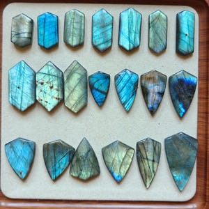 May include: A collection of twenty polished labradorite gemstones in various shield and hexagonal shapes. The stones display iridescent flashes of blue, green, and gold, with natural surface inclusions and veining visible across the smooth surfaces.