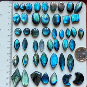 Può includere: Una collezione di pietre preziose di labradorite lucidata in varie forme, tra cui ovali, quadrati e gocce. Le pietre mostrano lampi iridescenti blu, verdi e dorati. È visibile un righello con marcature in centimetri.