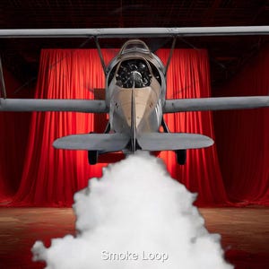 May include: A vintage biplane in flight, set against a backdrop of red curtains. The aircraft is silver and tan, with a cloud of white smoke billowing from beneath. The text "Smoke Loop" is visible at the bottom.