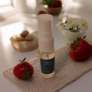 May include: A clear glass bottle of strawberry-scented oil with a white cap and a dark blue label. The bottle is next to two ripe strawberries and a small, beige linen cloth. The background includes a small bowl of strawberries and white flowers.