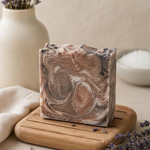 May include: A rectangular bar of soap featuring a marbled pattern of brown, black and white swirls. The soap has a textured, uneven top edge and sits on a square wooden soap dish with horizontal grooves, surrounded by scattered dried lavender sprigs.