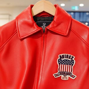 May include: A bright red leather jacket with a collared neckline and a zipper closure. The jacket features a patch on the chest with the text "AVIREX" and a design incorporating the American flag. The jacket is displayed on a wooden hanger.