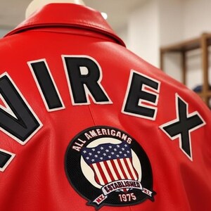 May include: Red leather jacket with the word "AVIREX" in large black letters outlined in white on the back. A circular patch with the American flag and the text "ALL AMERICANS ESTABLISHED 1975" is also visible.