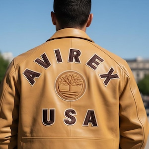 Może przedstawiać: Jasnobrązowa skórzana kurtka z napisem "AVIREX" w brązowych literach na plecach, wraz z logo drzewa i napisem "USA". Kurtka ma klasyczny kołnierz i dopasowany krój.