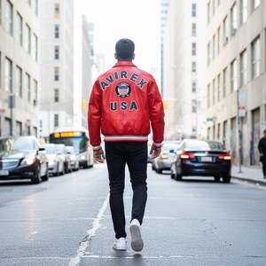 May include: A person wearing a red leather jacket with the text "AVIREX USA" on the back, walking down a city street. The jacket has white and blue accents. The person is wearing black pants and white sneakers.