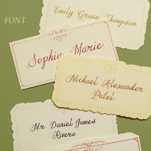 May include: A collection of personalized name cards with scalloped edges. The cards feature elegant calligraphy in various colors, including gold, red, and black. The names include "Emily Grace Thompson," "Sophia Marie," and "Michael Alexander Patel."