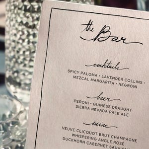 May include: A printed bar menu with the words "the Bar" in elegant script. The menu lists cocktails, beer, wine, and spirits, including Spicy Paloma, Peroni, and Grey Goose Vodka. The menu is on a white card with a black border.