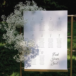 Puede incluir: Un plano de asientos de boda con un marco de metal dorado. El letrero blanco muestra los nombres de los invitados organizados por número de mesa. Delicadas flores blancas caen en cascada por el costado. El texto "Find Your Seat" está escrito en una elegante escritura.